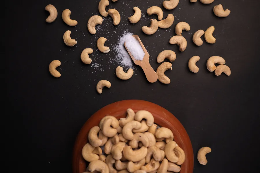 Salted cashews scattered around a clay bowl, with a wooden scoop of salt