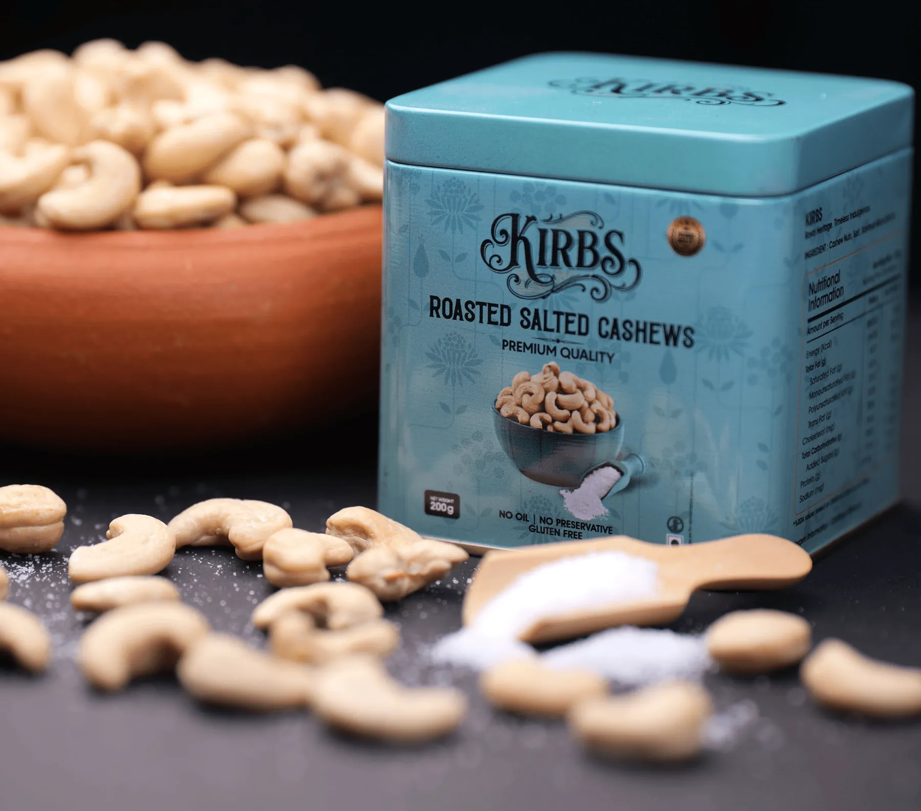 The teal Roasted Salted Cashews tin beside a clay bowl of cashews and a wooden scoop of salt